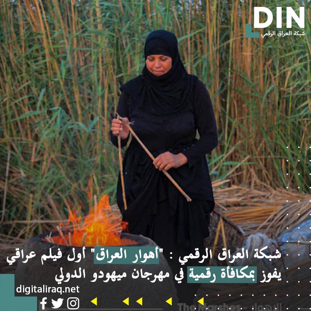 Woman in black hijab tending a small fire in a reed filled outdoor area, holding wooden sticks 